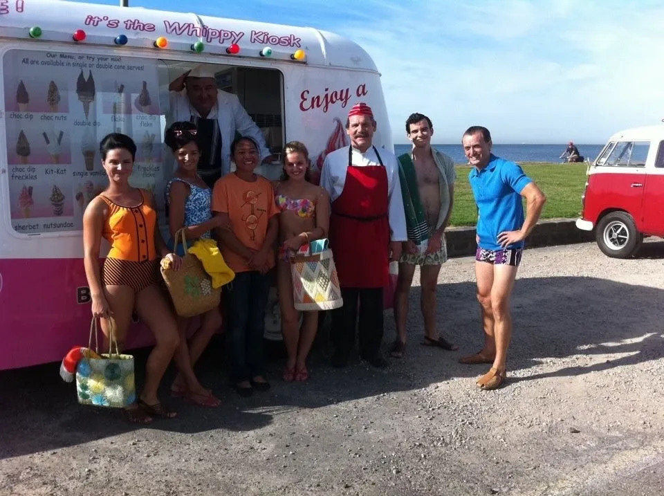 Cast and crew in period clothing standing beside the classic ice cream van during a production shoot
